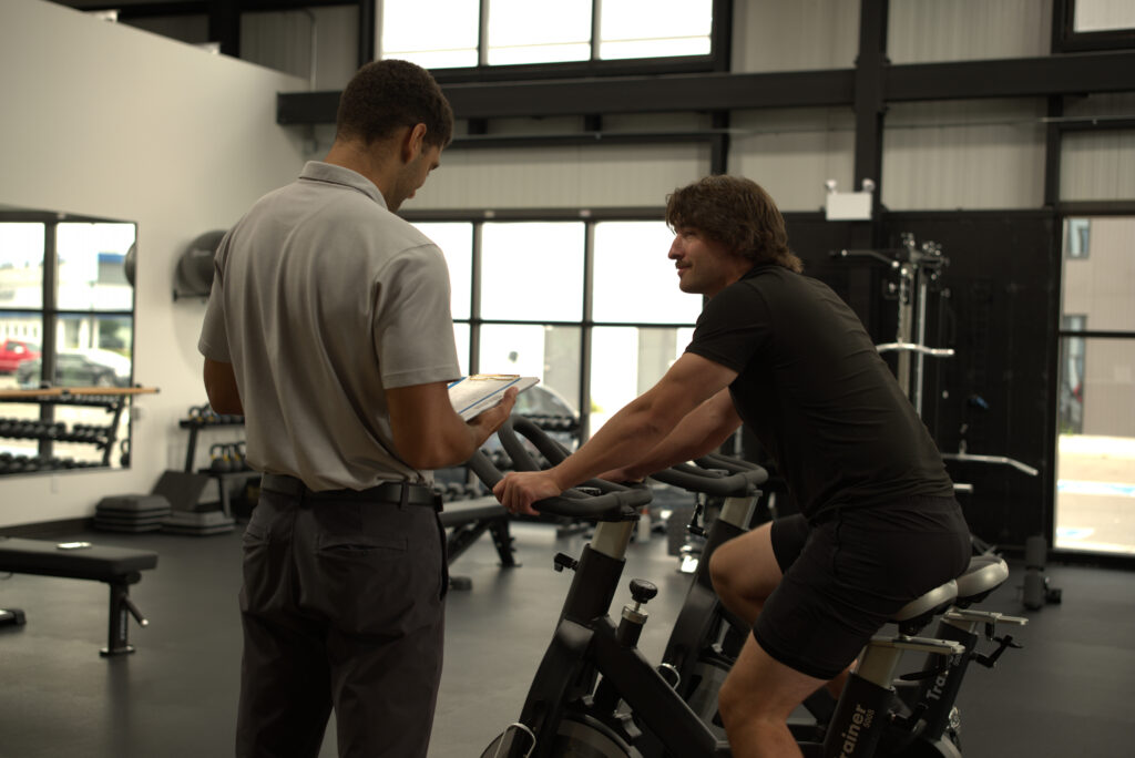 Dr Aden Cockburn guiding a patient through exercises on a stationary exercise bike to improve mobility and performance.